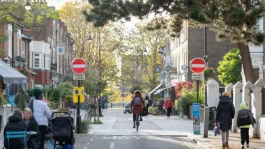 Person on bike on small road in London with people eating at outdoor dining tables and pedestrians walking on either side.